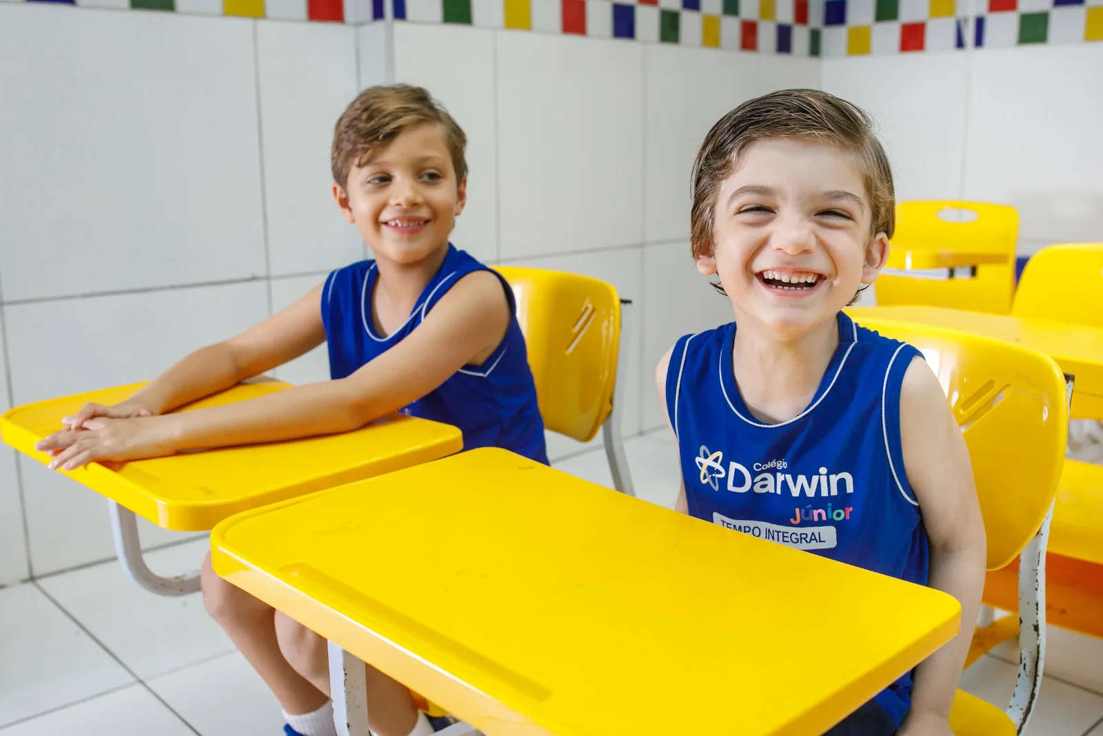 Dois alunos do Tempo Integral do Darwin, com a camiseta do STI, sorrindo nas carteiras amarelas da Sede Aldeota.